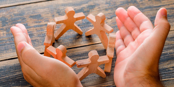 Manos protegiendo figuras humanas de madera como símbolo de cuidado, protección y apoyo comunitario.